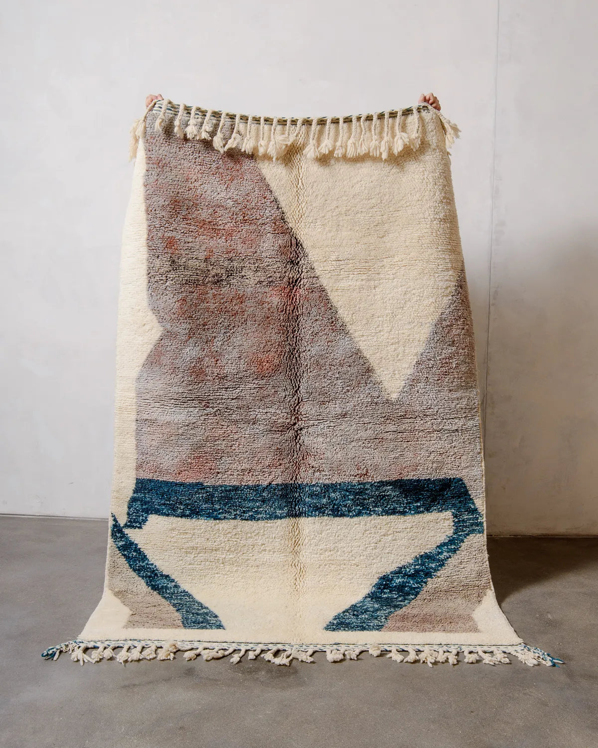 Wool Beniourain rug that is mostly cream and a mottled lavender-rust colour being held up in front of a white wall.
